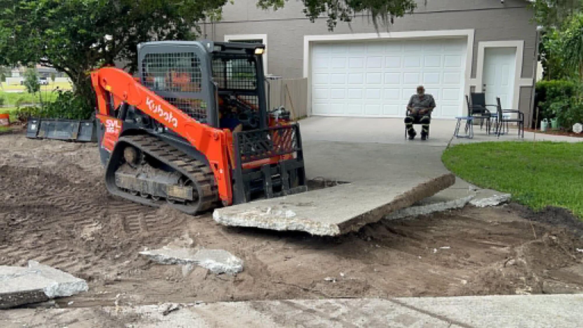 Skid steer Kubota de Javi's levantando una losa rota de una entrada de concreto demolida frente a un garaje