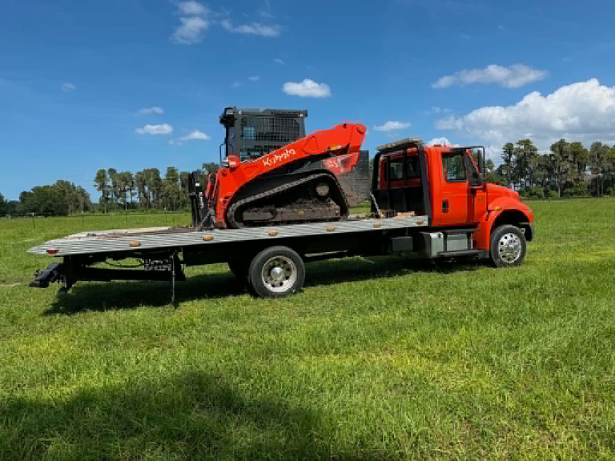 Rear view of Javi's rollback flatbed with a Kubota compact track loader chained down on the deck, palms and open field behind