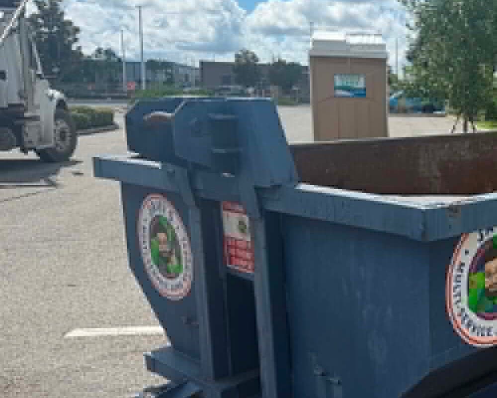Javi's empty 10 yard dumpster at the left of the frame in a commercial parking lot, the roll-off truck behind it